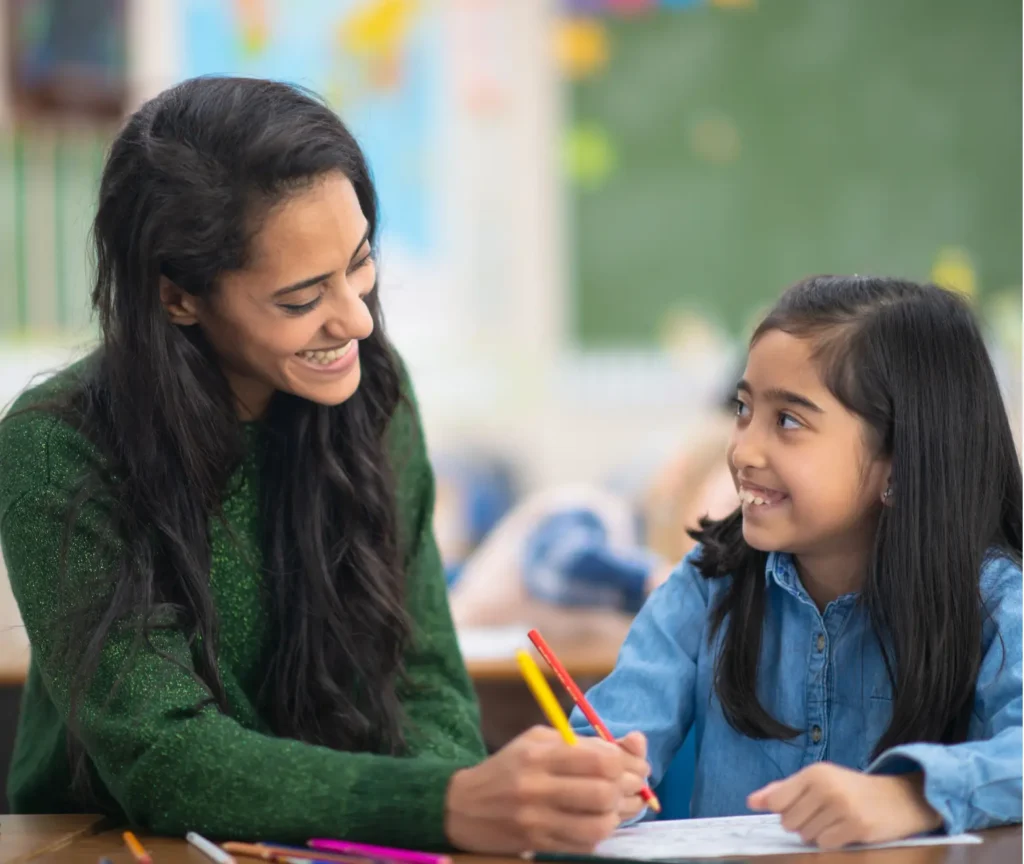 Teacher smiling and helping a young girl with her schoolwork at a classroom table