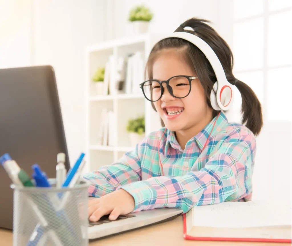 Smiling young girl wearing headphones and glasses while using a laptop at her desk