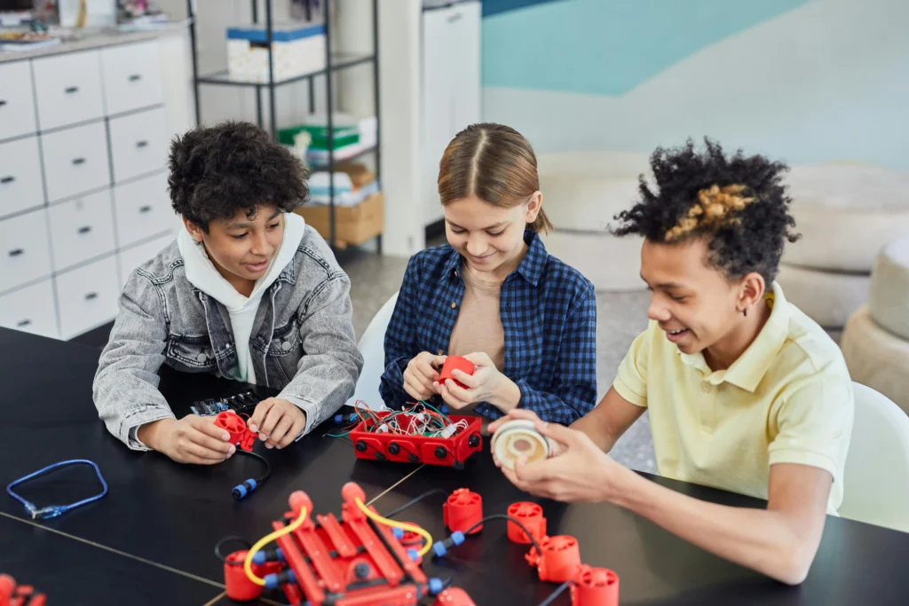Three students working together on a robotics project with red electronic parts at a classroom table.