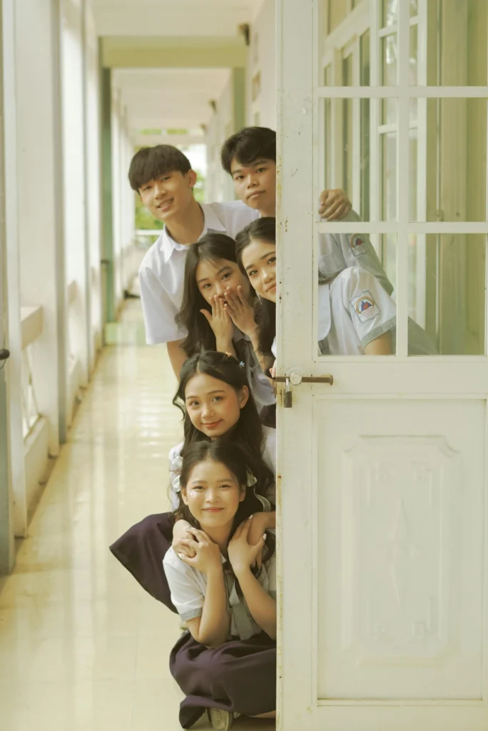 Group of smiling students in school uniforms peeking out from behind a door in a bright hallway.