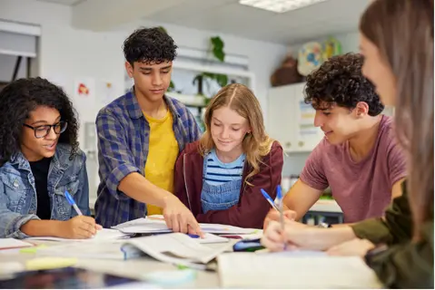 A group of teenagers working together at a table, discussing notes and pointing at a book.