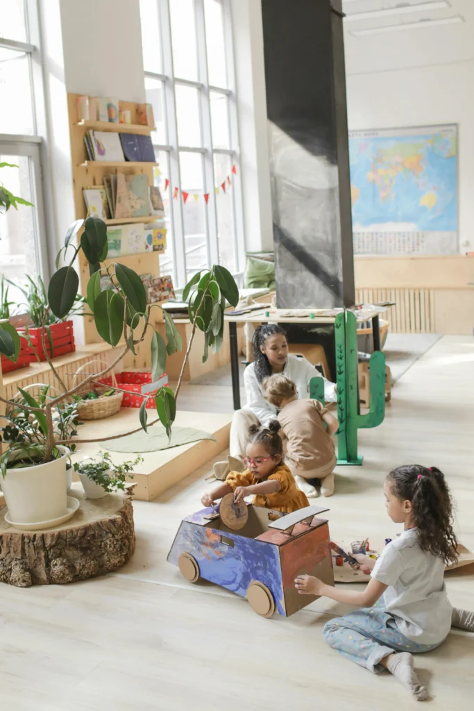 Children playing with cardboard toys in a bright, open classroom while an adult supports another child nearby.