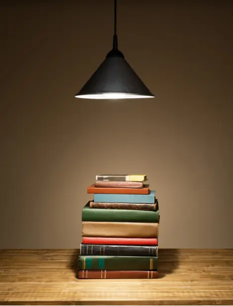 A stack of books on a wooden table under a single hanging lamp, creating a warm, focused study atmosphere.