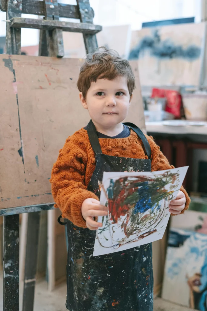 Young child holding a painting in an art classroom, showing learning through creative activity.
