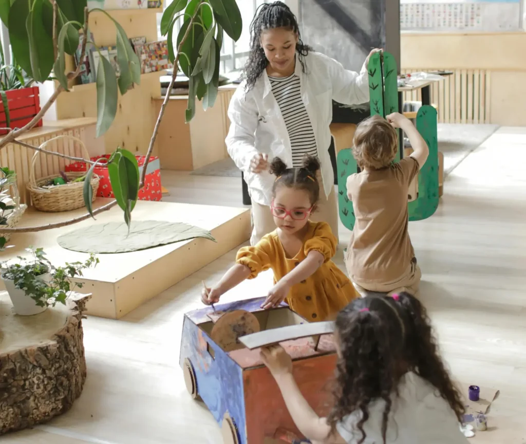 Children engaged in play-based learning in a classroom, creating and painting a cardboard project with guidance from a teacher.