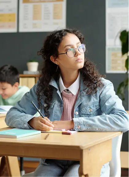 Adolescent student sitting at a desk in a classroom, writing and looking thoughtfully ahead.