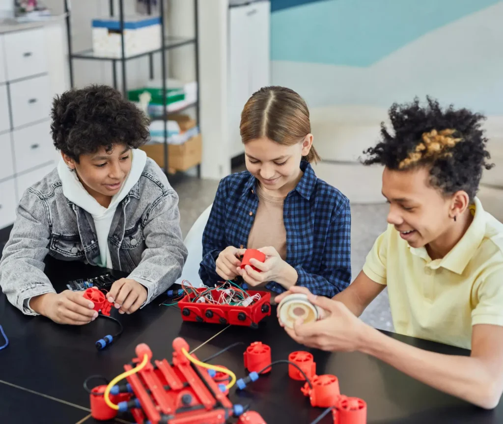 Three students working together on a robotics project with red electronic parts at a classroom table.