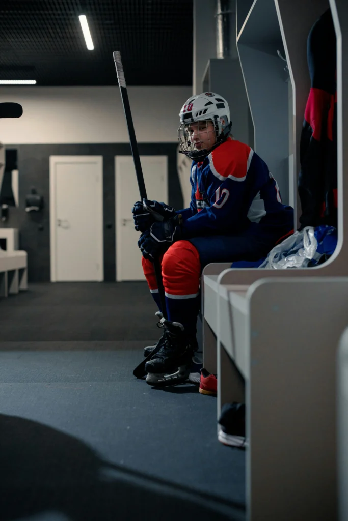Young ice hockey player sitting in a locker room holding a stick, reflecting before a game, representing youth sport development and early talent pathways.