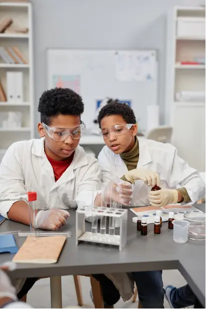 Two children wearing lab coats and safety glasses working together on a science experiment at a classroom table