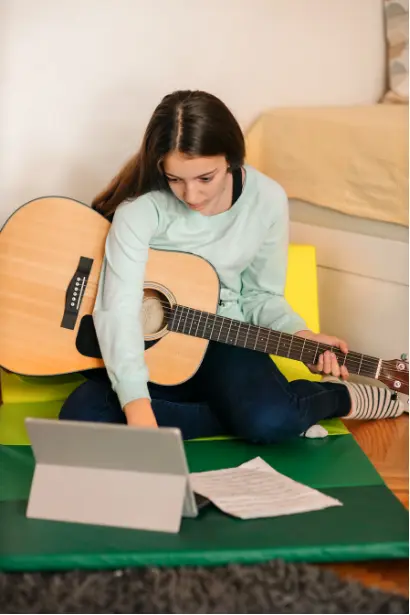 Teen girl practicing guitar while using a laptop for learning, combining creativity, focus, and cognitive development.
